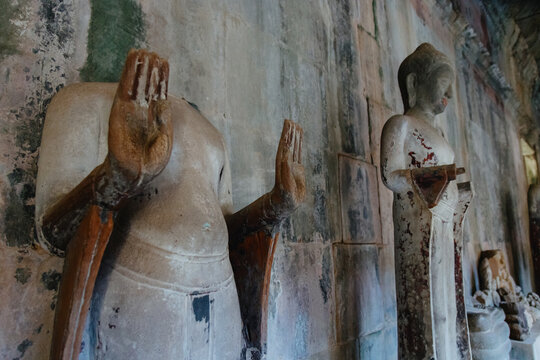 Ancient stone statues stand in a weathered temple hall in Siem Reap, Cambodia. The statues, some headless, show signs of age and decay.