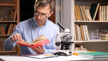 Young man wearing glasses intently reading a book, learning science in a laboratory setting with a microscope and bookshelves in the background, focusing on education and research © Aliaksandr