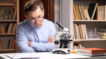 Man wearing glasses looking disappointedly at a microscope, experiencing failure during a scientific experiment in a laboratory setting with bookshelves in the background © Aliaksandr
