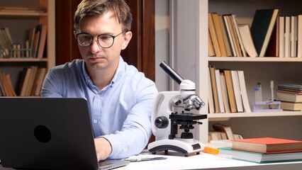Dedicated male scientist wearing glasses concentrating on his work, typing on a laptop with a microscope on the desk, surrounded by bookshelves in a laboratory setting © Aliaksandr