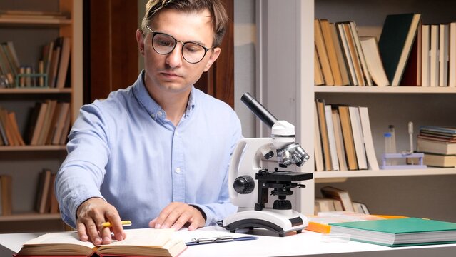 Male scientist wearing glasses studies an open book while using a microscope at a lab desk, surrounded by bookshelvesfocused on research, learning, and scientific discovery