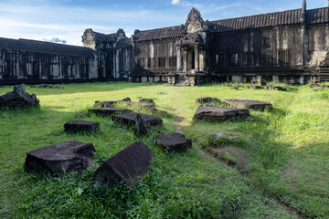 Fototapeta premium Angkor Wat, Siem Reap, Cambodia. Ancient stone ruins lie scattered on the grass in front of the temple complex, bathed in soft sunlight under a blue sky.