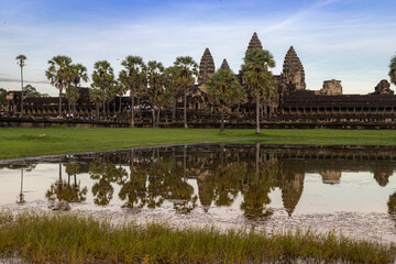 Fototapeta premium Angkor Wat, Siem Reap, Cambodia. Tourists admire the iconic temple complex at sunrise, reflected in the surrounding moat. The ancient Khmer architecture stands majestically against a soft sky.