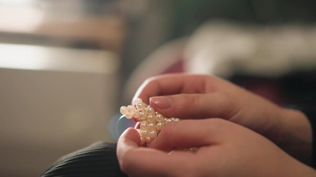 Close view of assembling delicate pearl jewelry. Detailed shot of skilled hands creating pearl necklace piece. Intimate portrayal of careful handwork in quiet jewelry workshop setting