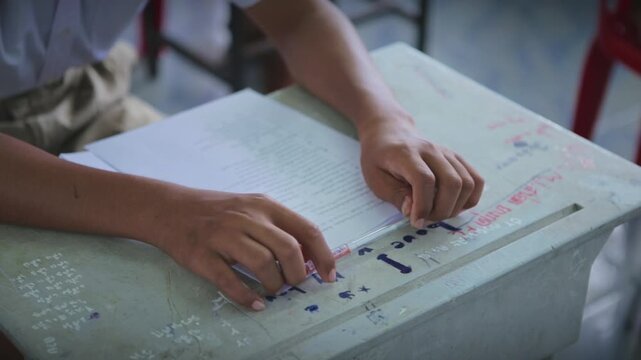 Close up of student hands holding a pen on desk with exam paper in classroom.