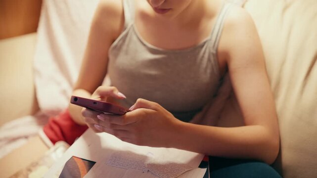 Focused high school girl sitting on a couch and typing on her mobile phone, using a smartphone to copy notes, browse exam answers, or cheat on a future test while studying at home.