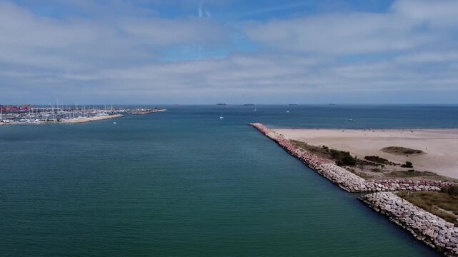 4K aerial drone video ascending and tilting down over the Turia river mouth in Valencia, Spain. The shot captures the port exit, a stone breakwater, sandy beach, boats, and Mediterranean Sea.