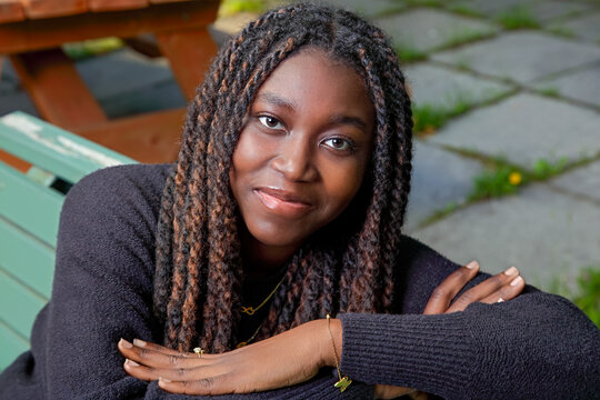 young African American woman with arms crossed and long braids resting on a green park bench smiling at the camera