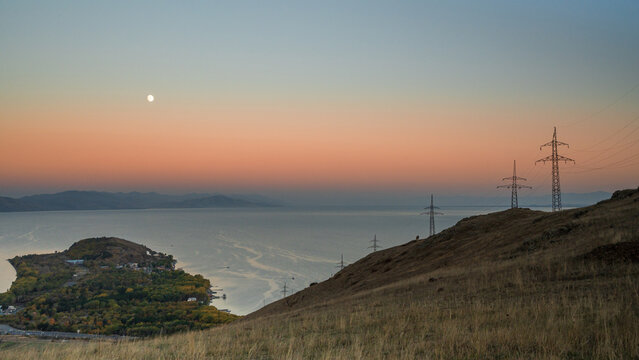A stunning panoramic view of Lake Sevan during twilight, showing the glowing moon above the peaceful water and the silhouette of hills in soft pastel hues.