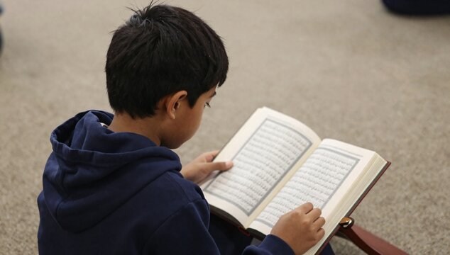 Young boy concentrates on reading the Quran on a wooden stand in an indoor setting