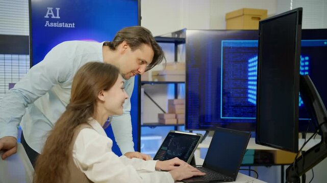 Male software developer points at monitor explaining code to female data scientist in modern AI research office. Team collaboration, AI development data analysis software engineering in tech industry.