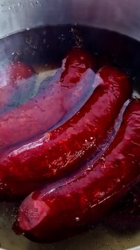 A close up shot of three red smoked sausages simmering in a stainless steel pot of boiling water. Authentic home cooking and food preparation scene.