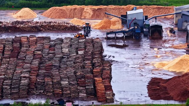 An aerial view of a large industrial sawmill featuring massive stacks of logs, wood chips, and active processing machinery with smoke stacks.