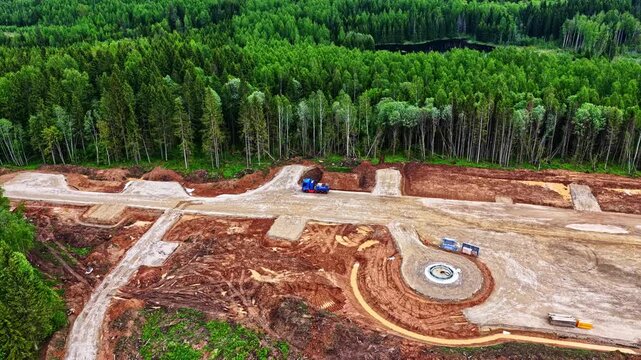 Cinematic aerial shot of the initial construction phase for a sustainable wind energy farm in Augstkalni, Latvia. Featuring heavy machinery, foundation work, and surrounding green forest.