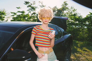 Young woman with retro style stands by a car in a sunny outdoor setting, wearing a striped rainbow...