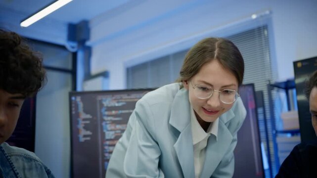 Female leader overseeing two male software developers working on laptops in modern corporate office. Project supervision, team management, workplace mentoring, and business oversight in tech company.
