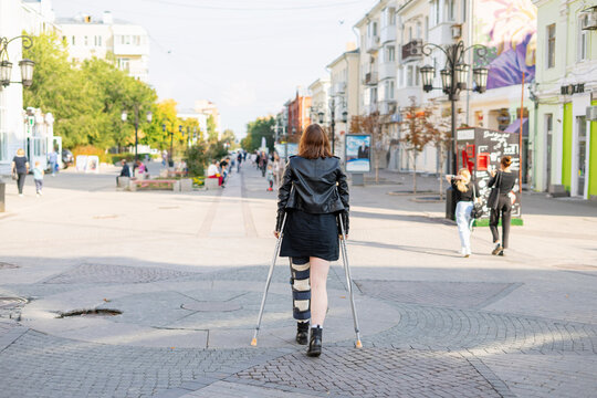 Young girl with a plastered foot in the orthosis and with crutches on a walk in the city