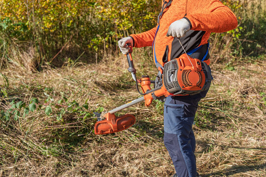 A man is using a string trimmer to cut weeds and grass in the field. He is wearing work clothes and gloves. Weed control and landscaping concept. Outdoor work, lawn care and.