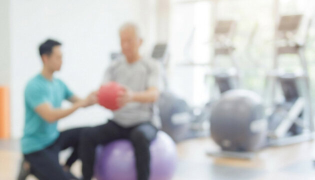 A trainer assists a senior individual in a gym, focusing on strength training with a medicine ball while seated on a stability ball in a bright environment.