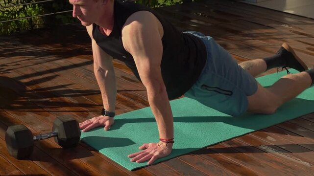Overhead perspective captures a fit young man with red hair transitioning from child's pose, sliding forward into a cobra stretch to extend his back on a green yoga mat outdoors. Static high angle med