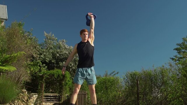 Fit young man with red hair lifts a kettlebell from the ground to his shoulder and presses it overhead on a lawn. Low angle wide view with a slight upward tilt to track the vertical movement.