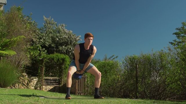 Fit young man with red hair executes full kettlebell swings on a lawn, training his glutes, hamstrings, and shoulders on a clear day. Static low angle wide view.
