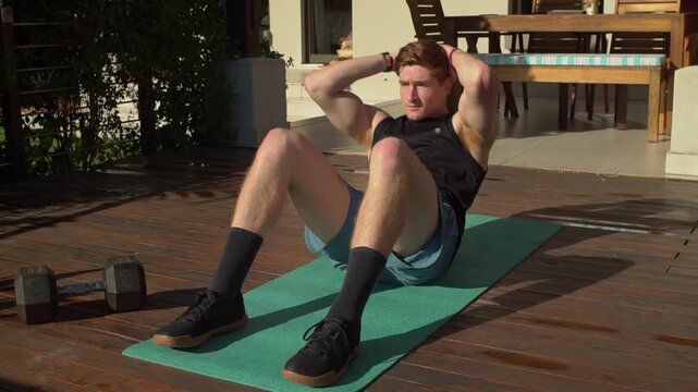Fit young man with red hair performs sit ups to strengthen his core and abdominal muscles on a green yoga mat over a wooden deck. High angle medium view.