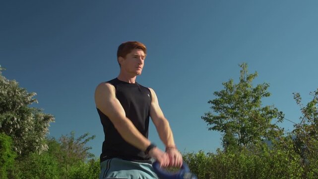 Fit young man with red hair thrusts his hips forward to complete kettlebell swings, training his shoulders and lower body on a clear day. Static low angle medium view.