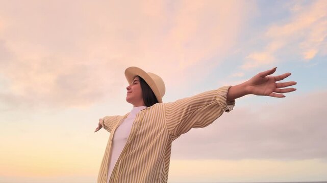 Young carefree woman in a hat enjoying freedom and life, breathing fresh air with open arms against a beautiful pastel sunset sky. Peaceful and relaxing moment during a summer vacation by the ocean