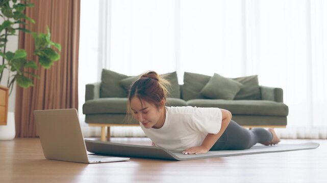 Young Asian woman stretching yoga workout on exercise mat while online training class with computer laptop in living room