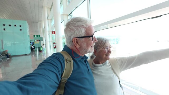Happy senior tourist couple with backpacks taking a selfie video inside an airport terminal, smiling and waving at the camera while waiting for their flight before starting their vacation