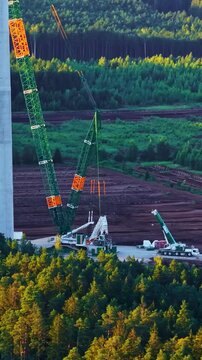 Aerial parallax over wind turbine construction site with massive crawler crane lifting tower section amid forest at golden sunset