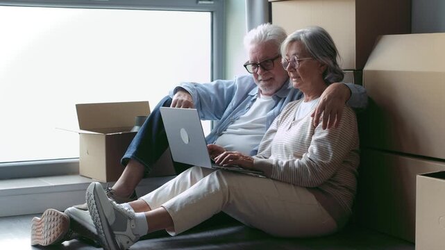Mature husband and wife sitting on the floor of their new house, surrounded by cardboard boxes, happily using a laptop together to browse for furniture or plan their next steps