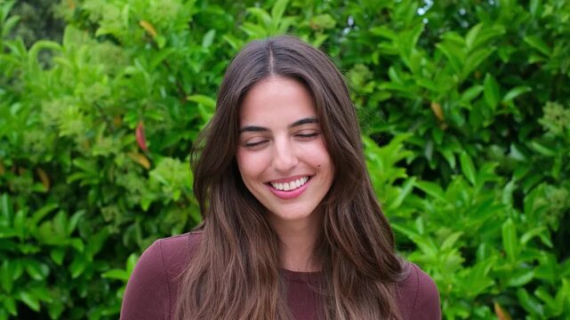 Portrait of an attractive young woman with long brown hair smiling genuinely at the camera while standing outdoors, with a lush green natural background creating a positive and serene mood