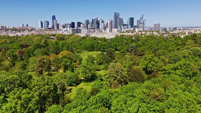 Drone view over green expanse of Bois de Boulogne with modern skyscrapers of La D&eacute;fense business district in background in Paris, France. Aerial forward ascending