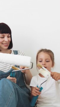 A mother and her young daughter sit on the floor, holding paint rollers and preparing to redecorate a room
