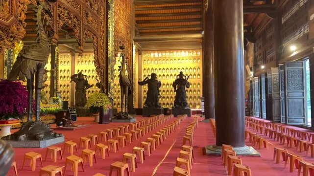 Wide view inside Bai Dinh Pagoda in Ninh Binh, Vietnam showing bronze guardian statues, golden Buddha, carved wooden pillars, red carpet and prayer stools