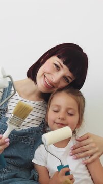 A mother and her young daughter playfully hold paintbrushes and rollers against a plain white background, enjoying a home renovation project together