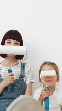 A mother and her young daughter playfully hold paint rollers in front of their faces while preparing to redecorate a room