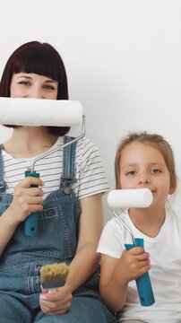 A mother and her young daughter playfully hold paint rollers in front of their faces while preparing to redecorate a room