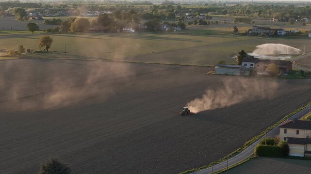 Tractor performing soil tillage across cultivated field with irrigation systems and rural settlements revealed during upward camera tilt at sunset in Po Valley Italy slow motion parallax aerial shot