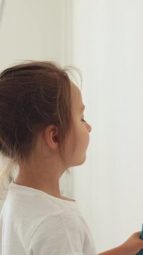 A young girl with blonde hair holds a paint roller and applies paint to a white wall while a ladder stands in the background