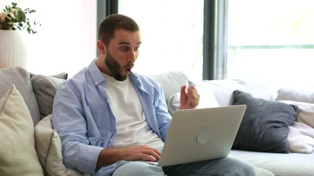 Excited young man sitting on the sofa at home, working on his laptop, and celebrating a successful achievement or winning a game with a triumphant gesture of happiness and joy