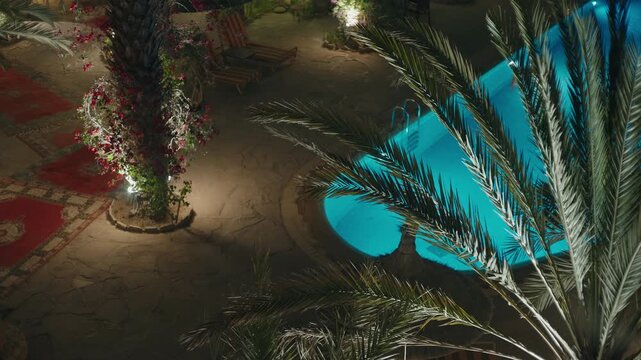 Overhead view of a Moroccan riad courtyard at night with glowing blue pool palm trees bougainvillea and red rugs
