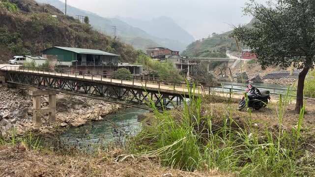 Metal truss bridge crossing Nho Que River near small village with mountain slopes local buildings and riverside vegetation in Ha Giang Vietnam