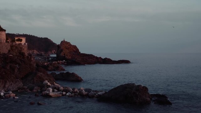 Moody twilight view of rugged coastline and sea stack near Riomaggiore in Cinque Terre Italy. Dark cliffs rise beside the calm Mediterranean waters