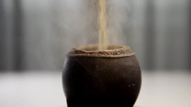 Close view of loose tea or dried herbs pouring into traditional dark ceramic vessel. Fine particles fall steadily against the soft blurred background