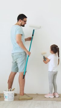 A father and his young daughter are painting a white wall together with paint rollers, enjoying a home improvement project
