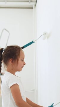 A father and his young daughter are painting a white wall together, using rollers and extension poles