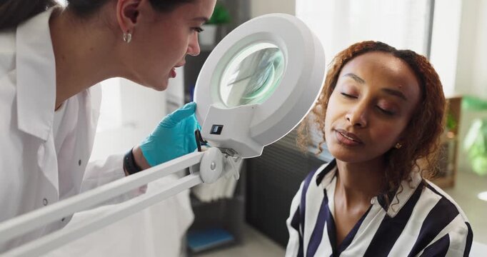 Dermatologist doctor examines patient skin in clinic. A magnifying lamp supports examination of facial skin for acne, moles and pigmentation. Clear trusted healthcare and consultation concept.
